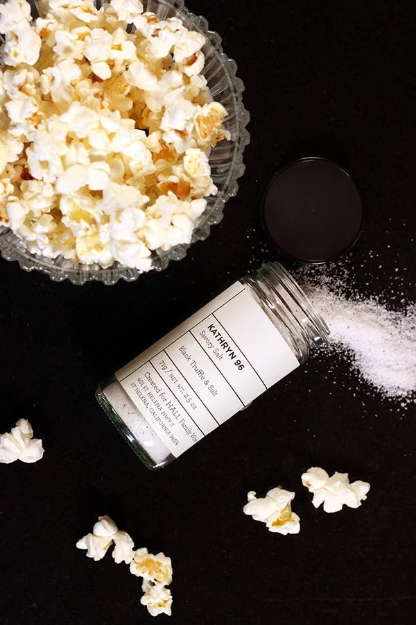Jar of HALL Kathryn 06 Black Truffle Salt laying flat on a black background next to a bowl of popcorn.